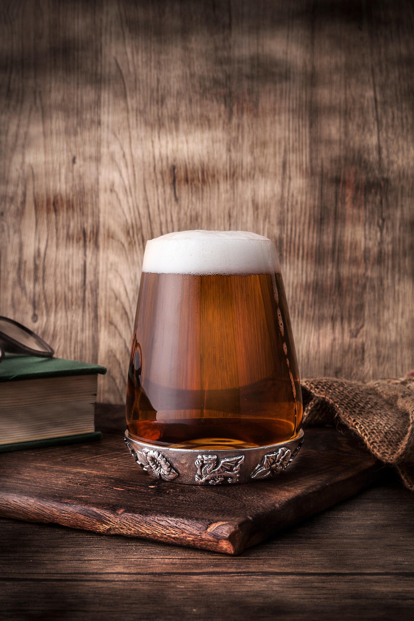 Glass tumbler with detailed Welsh dragon pewter base design sitting on an oak board in a rustic scene