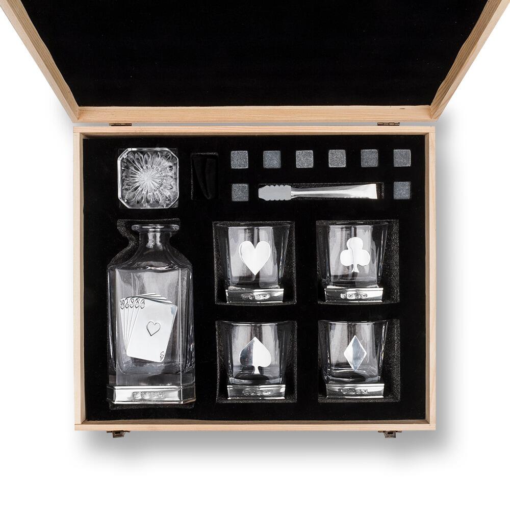 Overhead view of an opened wooden gift box containing a whiskey decanter with playing card design, four glasses with card suit emblems, whiskey stones, and metal tongs, all arranged neatly in black foam padding"