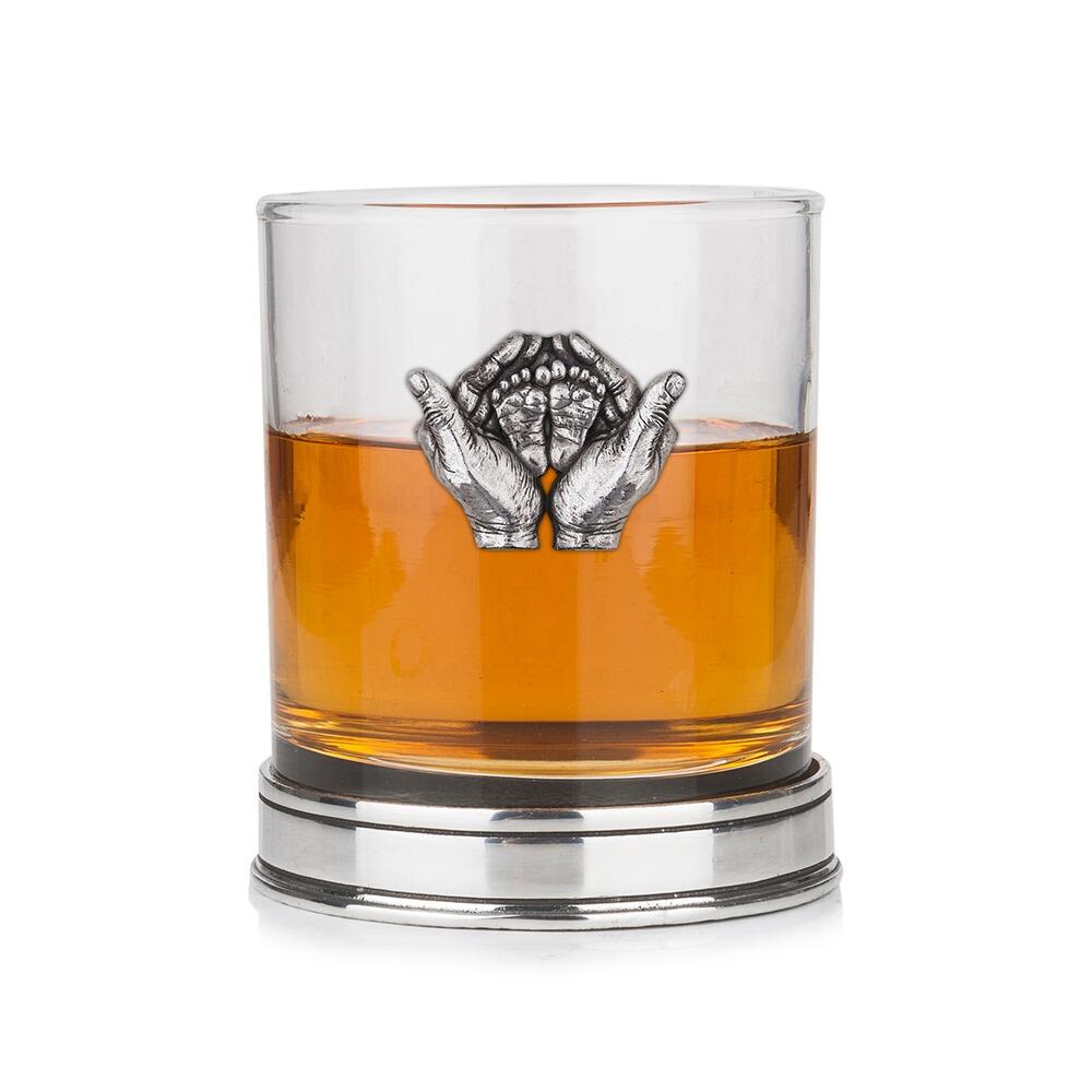 Whisky tumbler with pewter hands holding baby feet emblem and polished base, partially filled with amber liquid
