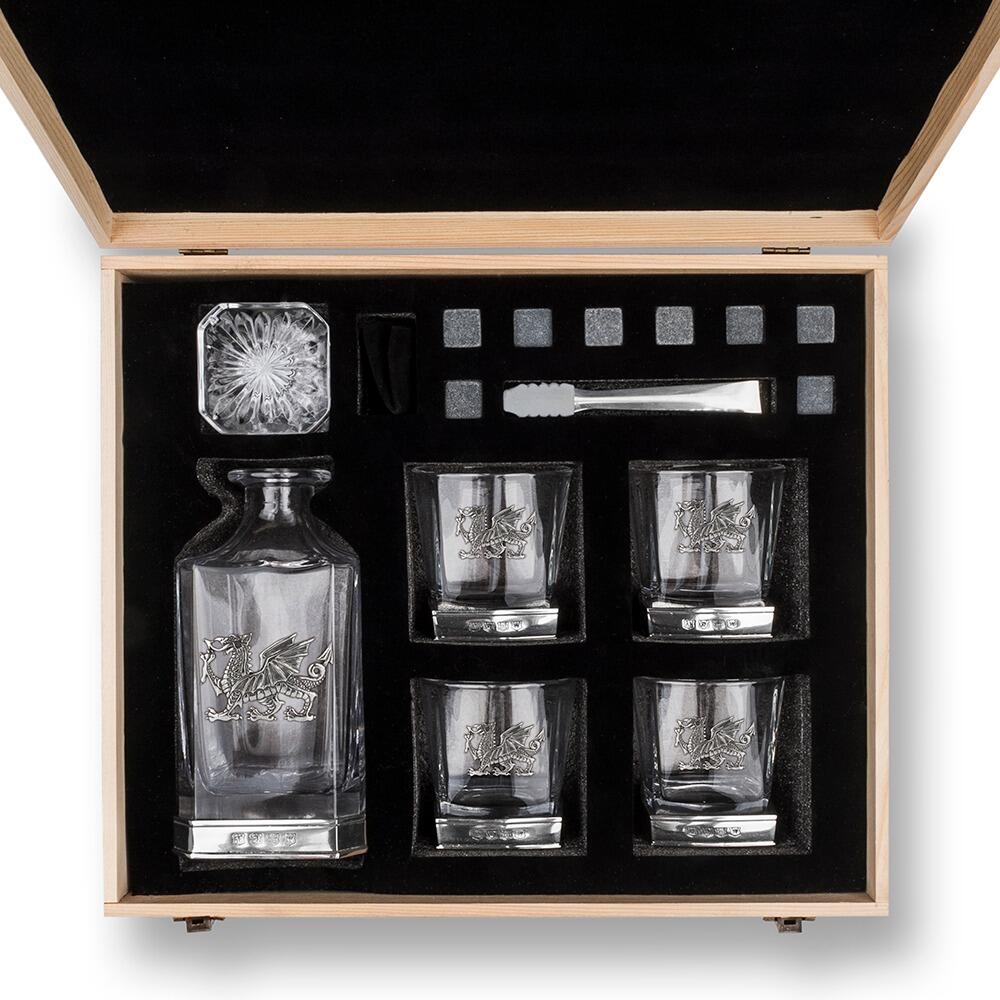 Overhead view of an opened wooden gift box containing a whiskey decanter with a Welsh Dragon, four glasses with Welsh Dragon motifs, whiskey stones, and metal tongs, all arranged neatly in black foam padding