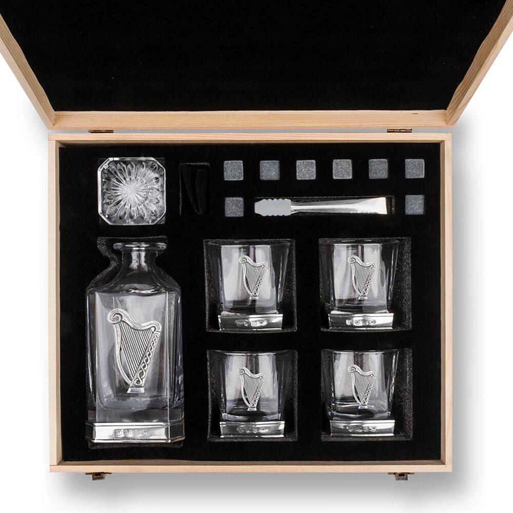 Overhead view of an opened wooden gift box containing a whiskey decanter with Harp, four glasses with Harp motifs, whiskey stones, and metal tongs, all arranged neatly in black foam padding"