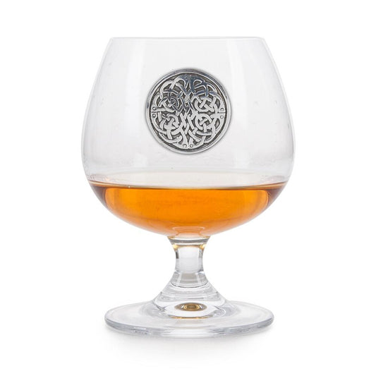 Crystal brandy glass with pewter Celtic Never Ending Knot emblem, partially filled with amber liquid