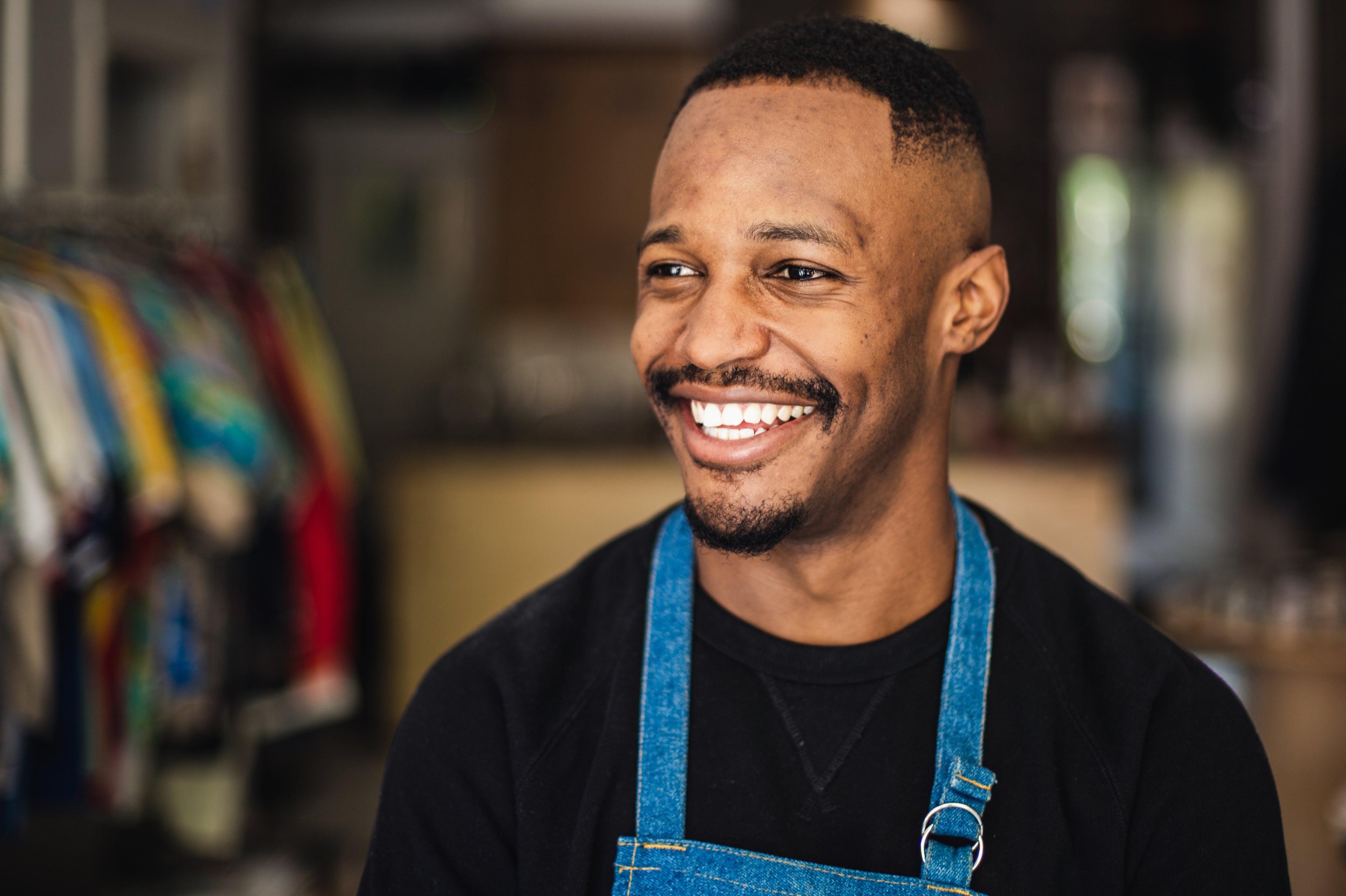 Smiling man wearing a black shirt and denim apron in a colorful clothing store.