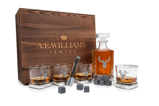 Wooden presentation box displaying decanter and four tumblers with pewter stag motifs, accompanied by whisky stones and tongs
