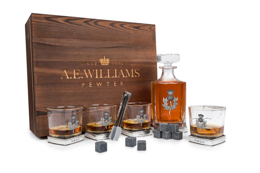 Wooden presentation box displaying decanter and four tumblers with pewter thistle motifs, accompanied by whisky stones and tongs