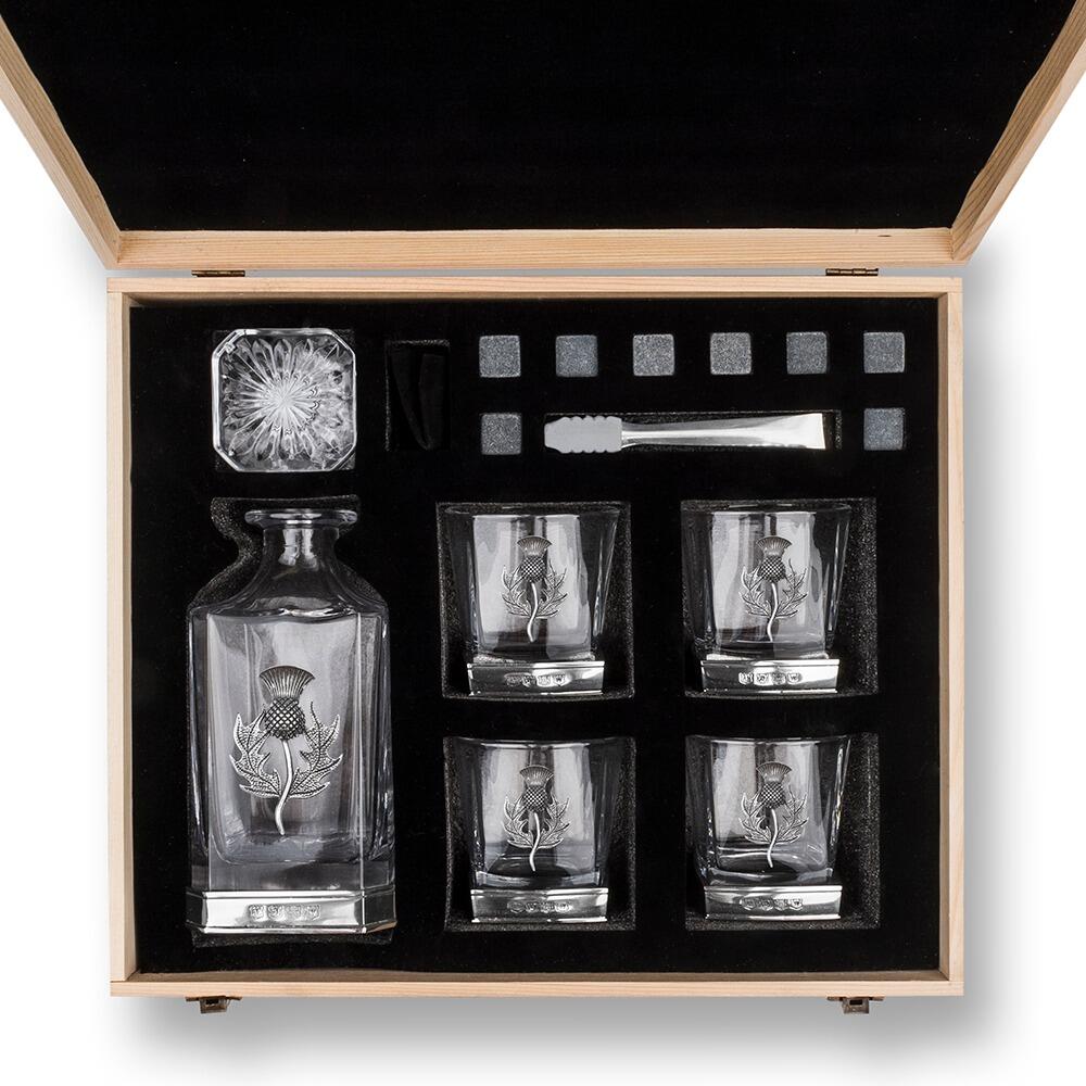Overhead view of an opened wooden gift box containing a whiskey decanter with Thistle, four glasses with Stag motifs, whiskey stones, and metal tongs, all arranged neatly in black foam padding