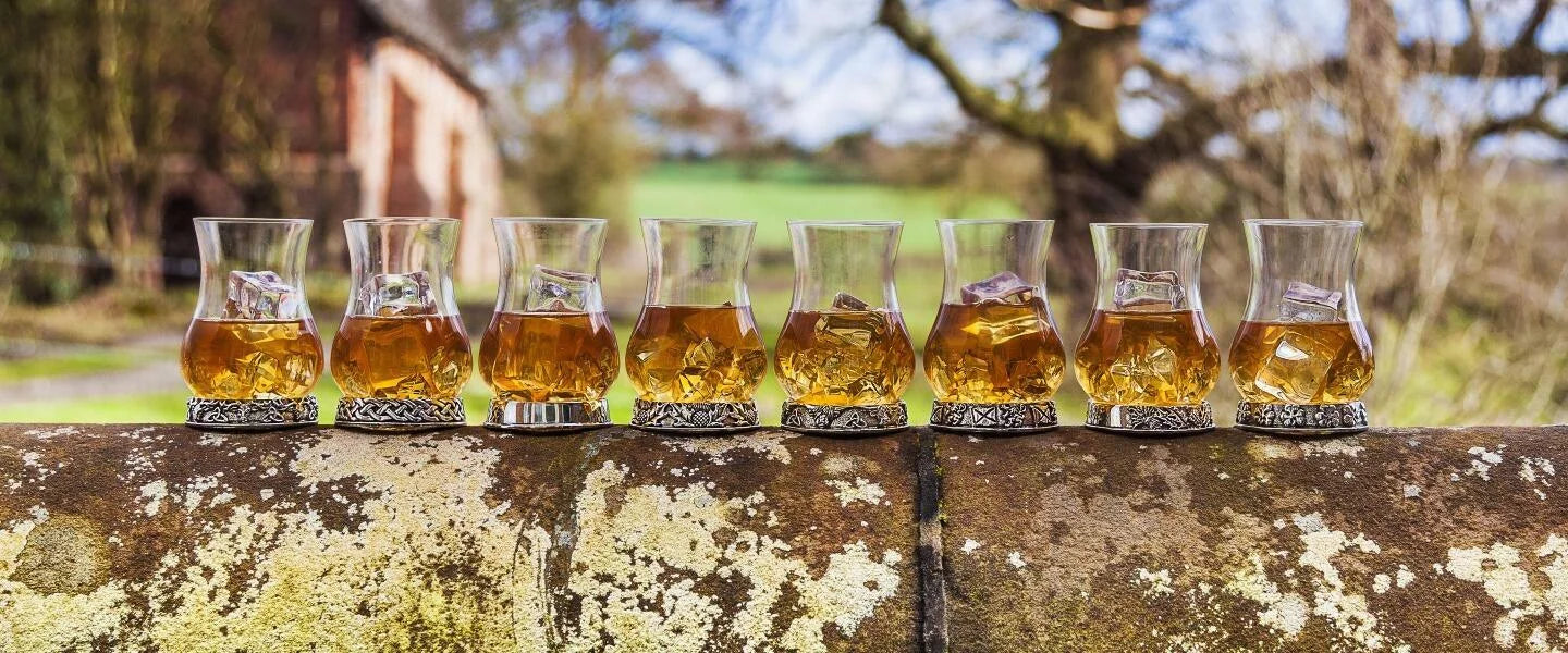 Set of eight whiskey glasses with silver bases, filled with amber whiskey and ice, outdoors with a scenic background.